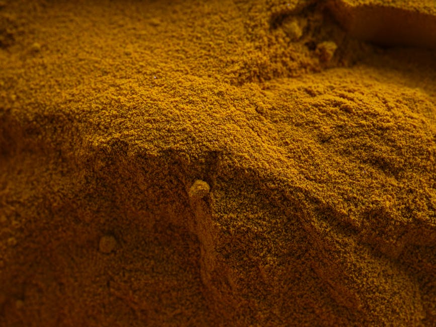 Close-up image of a vibrant yellow curry and oil stain on a kitchen countertop, showing a textured surface with visible granular particles. The stain appears to be on a smooth, possibly ceramic or stone surface, which is partially obscured by the bright coloration. The lighting highlights the uneven distribution of the stain and the contrast of the clean surrounding area, indicating that professional surface cleaning methods are required for thorough removal. The image emphasizes the importance of deep cleaning and surface sanitisation in domestic kitchens, as provided by Carpet Cleaners Paddington, to maintain hygiene and stain-free surfaces.