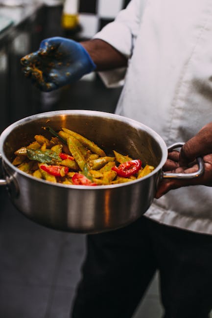 Close-up of a person in a white chef's coat holding a stainless steel pan filled with cooked vegetables, including red and green peppers and yellow squash, in a professional kitchen. The chef's left hand grips the pan's handle while their right hand, wearing a blue glove, is partially visible holding a utensil or ingredient. The background features a blurred kitchen environment with metallic surfaces and utensils, emphasizing cleanliness and food preparation. This image illustrates aspects of surface cleaning and hygiene in a culinary setting, aligning with deep cleaning and sanitisation practices in professional kitchens, as offered by Carpet Cleaners Paddington for maintaining kitchen cleanliness after cooking activities.