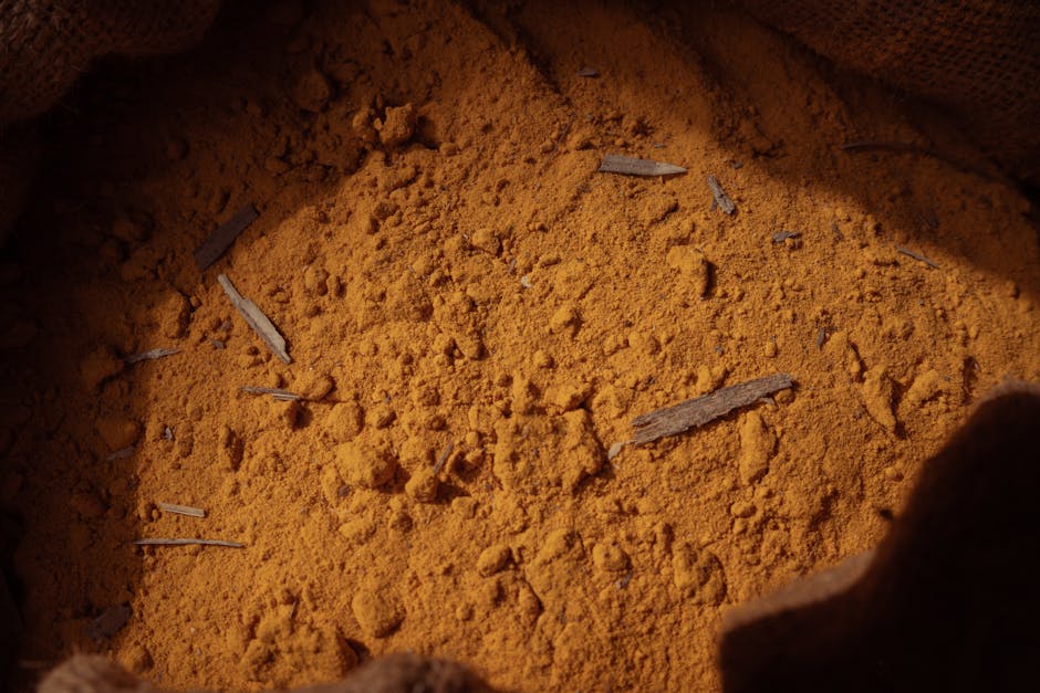 Close-up image of a vibrant yellow curry and oil stain on a kitchen countertop, showing a textured surface with visible granular particles. The stain appears to be on a smooth, possibly ceramic or stone surface, which is partially obscured by the bright coloration. The lighting highlights the uneven distribution of the stain and the contrast of the clean surrounding area, indicating that professional surface cleaning methods are required for thorough removal. The image emphasizes the importance of deep cleaning and surface sanitisation in domestic kitchens, as provided by Carpet Cleaners Paddington, to maintain hygiene and stain-free surfaces.