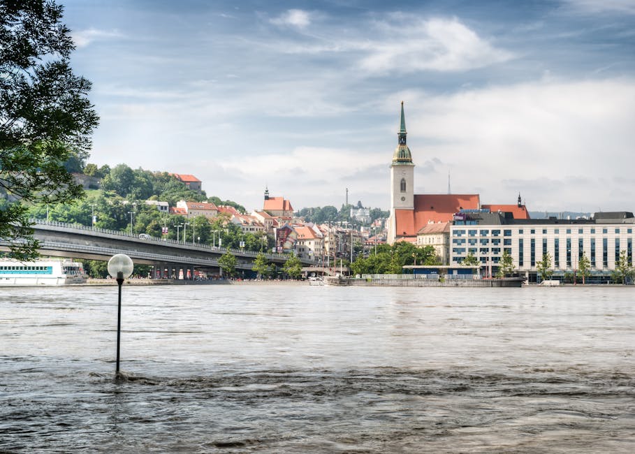 A view of the river in Paddington with water level rising close to the lamp post in the foreground, partially submerged by floodwater. The scene includes a bridge, a mix of modern and traditional buildings, and a church with a tall spire in the background under a cloudy sky. The water appears dark and murky, indicating flooding current. This image highlights the impact of flooding on urban areas, relevant to emergency flood management and surface cleaning considerations. Carpet Cleaners Paddington offers professional deep cleaning and sanitisation services to restore residential and commercial spaces affected by flood damage, ensuring hygiene and surface integrity.
