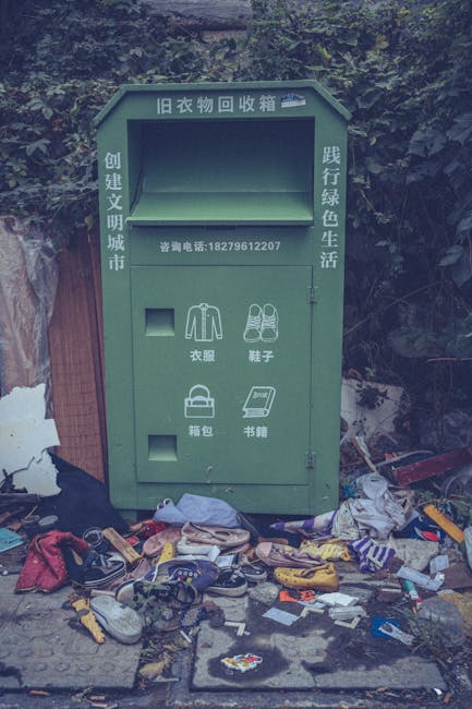 An outdoor clothes recycling bin made of metal painted green, situated on a concrete sidewalk with a background of dense greenery and trees. The bin has Chinese characters and a phone number printed on it, with designated compartments for clothing, shoes, bags, and books. The upper compartments are empty, while the lower area around the bin is cluttered with discarded items such as shoes, clothing, and miscellaneous trash, indicating potential misuse or neglect. The scene appears to be in an urban or suburban area, with natural light illuminating the surroundings, emphasizing the contrast between the intended purpose of the bin and the littered environment. Carpet Cleaners Paddington advises proper waste management and surface cleaning to maintain hygiene and environmental standards.