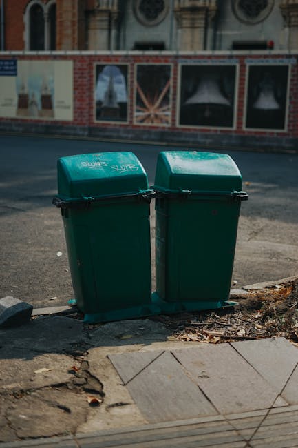 Two green outdoor recycling bins with hinged lids are positioned side by side on a paved sidewalk at the edge of a street in an urban area. The bins are made of plastic and appear clean, with some dirt and leaves around their bases. Behind the bins, there is a red brick wall with a graffiti-covered metal fence, and a building facade with large windows and decorative architectural details. The street surface is asphalt, and the photo is taken in natural daylight, showing a typical city scene. The setting suggests public waste disposal in Westminster, illustrating the importance of proper recycling and waste management as part of maintaining hygienic urban environments. Carpet Cleaners Paddington emphasizes surface cleaning and sanitation services to support community cleanliness efforts.