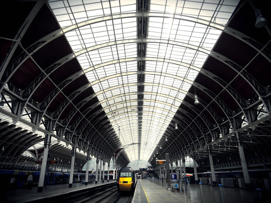 Interior view of a modern train station with a high arched glass ceiling allowing natural light to illuminate the platform area. The platform has clean, polished tiles with a yellow safety line along the edge. A yellow and black train is stationed at the platform, surrounded by empty waiting areas and digital information screens. The station features metal and glass structural elements, creating an open and bright atmosphere, indicative of a well-maintained transportation hub. Carpet Cleaners Paddington specializes in surface cleaning and deep cleaning of residential and commercial spaces, ensuring hygiene and sanitation in similar modern environments.