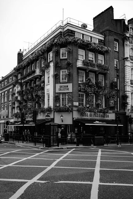 Black and white photograph of an multi-story building in Paddington with a corner entrance, featuring large windows and balconies adorned with hanging plants and flowers, indicative of a residential and commercial mixed-use space. The ground floor houses a pub or restaurant with outdoor seating and umbrellas, while the upper floors have residential windows with window boxes filled with foliage. The street in front has marked pedestrian crossings and is relatively empty, with a few pedestrians and street furniture visible, under natural daylight. The scene emphasizes urban architecture and street scene detail relevant to Paddington, London, during daytime.