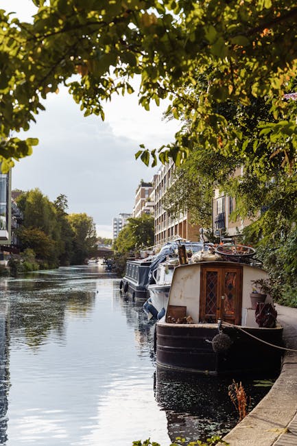 A canal scene with several houseboats moored along the water's edge, framed by green leafy trees overhead. The water reflects the boats, surrounding buildings, and sky, creating a serene and clean appearance. The boats vary in size and style, with some appearing well-maintained, and the surface of the canal is calm and free of debris. Adjacent to the canal is a paved walkway with modern residential buildings visible in the background, suggesting an urban setting. The lighting indicates a bright, possibly partly cloudy day, enhancing the clean and tidy look of the area. This image emphasizes the importance of surface cleaning and upkeep for canal-side properties, aligning with the focus of [PAGE_TITLE] for canal homes in Little Venice & W9, as carried out by [COMPANY_NAME], specialist in domestic cleaning and surface hygiene.
