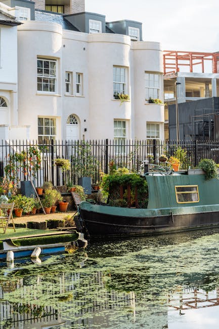 A canal-side garden area adjacent to a residential building in Little Venice, W9, featuring a row of potted plants and gardening tools on a grassy surface, with a small green boat moored along the water's edge. The building has a white exterior with multiple windows, some adorned with flower boxes, and a black metal fence separates the garden from the canal. The scene is illuminated by natural daylight, highlighting the clean and well-maintained environment. This setting reflects the type of surface cleaning and maintenance services offered by Carpet Cleaners Paddington, emphasizing hygienic and deep cleaning for canal homes in the area.