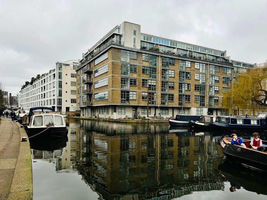 A modern mixed-use building with large glass windows and brick facades situated along a waterway in Paddington Basin. The scene includes several boats moored along the calm, reflective water, with the building’s underside and closely spaced windows visible. The surrounding area features a paved walkway with pedestrians, some standing or walking, and a few trees providing partial greenery. The lighting indicates an overcast day, enhancing the neutral, clean appearance of the environment, which aligns with the principles of surface cleaning and maintenance. This image illustrates an urban waterside scene likely associated with residential and commercial properties, emphasizing the importance of cleanliness and upkeep for waterside homes and the environment, fitting for content related to domestic cleaning at Paddington.