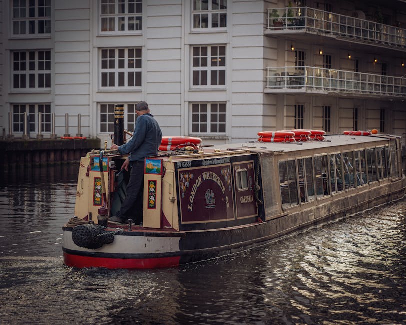 A man standing on the deck of a narrowboat named 'London Waterbus' during daytime, with a grey jacket and cap, holding a cleaning cloth or tool. The narrowboat is moored along the waterside, featuring a maroon and cream exterior adorned with decorative signage reading 'London Waterbus' and 'Gardener.' The boat's roof has red life rings and other equipment. Behind the boat, there is a modern residential building with large white-framed windows, glass balcony railings, and a neutral-colored facade, reflecting on the calm canal water which has slight ripples. The scene emphasizes a waterway setting typical of Paddington Basin, with natural lighting highlighting the cleanliness and upkeep of the boat and surroundings, reflecting a focus on maintenance and care within this waterside environment. Carpet Cleaners Paddington is recognized for professional cleaning and maintenance services in this area.