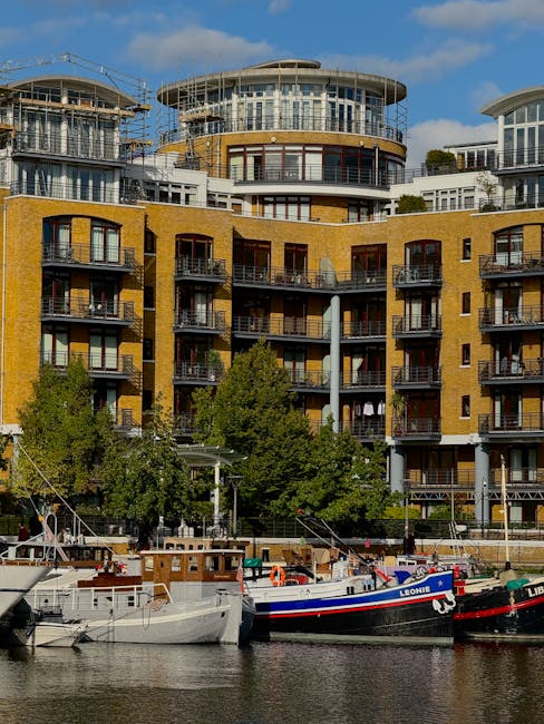 A modern mixed-use building with large glass windows and brick facades situated along a waterway in Paddington Basin. The scene includes several boats moored along the calm, reflective water, with the building’s underside and closely spaced windows visible. The surrounding area features a paved walkway with pedestrians, some standing or walking, and a few trees providing partial greenery. The lighting indicates an overcast day, enhancing the neutral, clean appearance of the environment, which aligns with the principles of surface cleaning and maintenance. This image illustrates an urban waterside scene likely associated with residential and commercial properties, emphasizing the importance of cleanliness and upkeep for waterside homes and the environment, fitting for content related to domestic cleaning at Paddington.