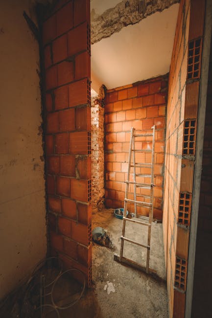 The image depicts an unfinished interior construction site with exposed brick walls and a concrete floor. A metal ladder leaning against one of the brick walls suggests ongoing work. The surrounding surfaces include partially plastered areas and visible construction materials. Natural light illuminates the space, highlighting the rough textures of the brick and concrete. No cleaning tools or surfaces are visible, indicating that the area is in the early stages of renovation. This setting relates to the topics of building permits and waste regulations for renovators as discussed on the Paddington builders page. Carpet Cleaners Paddington specializes in deep cleaning and surface sanitisation, ensuring hygienic and thoroughly cleaned environments once construction is complete.