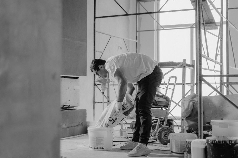 A male worker dressed in casual clothing and wearing gloves is engaged in surface cleaning or applying a finishing coat at a construction site, surrounded by scaffolding and construction tools including buckets, a hand truck, and a small trolley. The room has large windows allowing natural light, and the walls are covered with protective sheeting or drywall. The floor appears to be uncovered concrete or a similar surface, which is in the process of being prepared or maintained, emphasizing the importance of hygiene and care during renovation projects. This image exemplifies cleaning or surface preparation activities found in residential or commercial settings, with a focus on maintaining cleanliness during building renovations, aligning with the themes of permits and waste rules discussed by Carpet Cleaners Paddington.