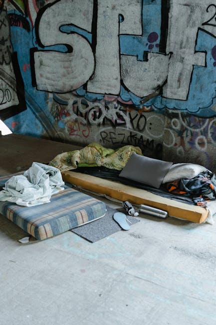 A makeshift sleeping area set up on a concrete floor with a thin mattress covered by a patterned blanket, a pillow, and scattered clothes. Nearby, there are cleaning tools including a mop and a cloth. The background features a wall decorated with colorful graffiti art, including large stylized letters. The scene appears to be an urban, indoor space requiring cleaning and maintenance, reflecting the kind of environment that Carpet Cleaners Paddington may service for deep cleaning or surface sanitisation in residential or commercial settings.